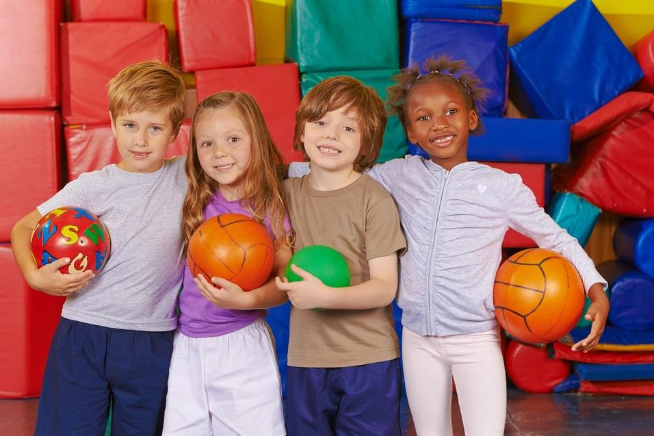 Vier Kinder halten bunte Bälle in einer Turnhalle.