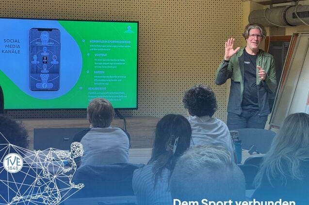 Eine Person steht vor einem Bildschirm und hält eine Präsentation, während eine Gruppe von Zuhörern im Hintergrund aufmerksam zuhört.