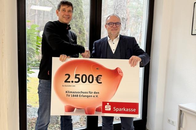 Zwei Personen stehen nebeneinander und halten einen großen Scheck mit einem Betrag von 2.500 € von der Sparkasse.