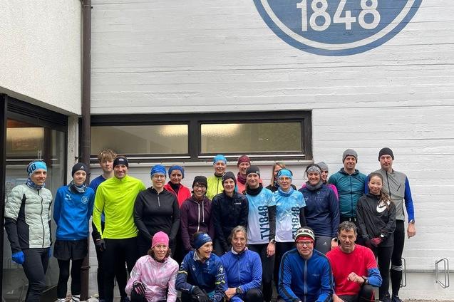Gruppe von Menschen in Sportkleidung versammelt vor der Wand des Hauptgeb&auml;udes des TV 1848 Erlangen mit gro&szlig;em Vereinslogo beim Community Run.