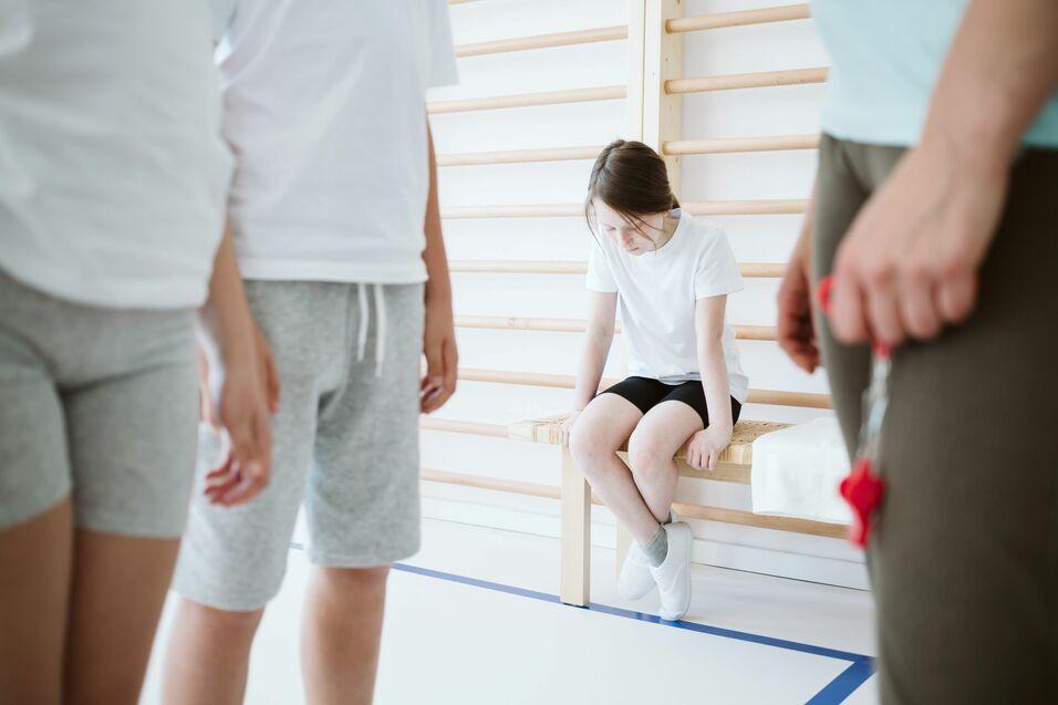 Prävention Gewalt Ein Mädchen sitzt mit gesenktem Kopf auf einer Bank in der Turnhalle, während andere Kinder und eine Person mit Trillerpfeife im Vordergrund stehen.