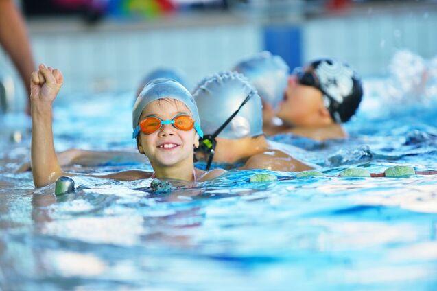 Ein Kind mit Schwimmbrille und Badekappe hebt lachend eine Faust im Schwimmbecken.